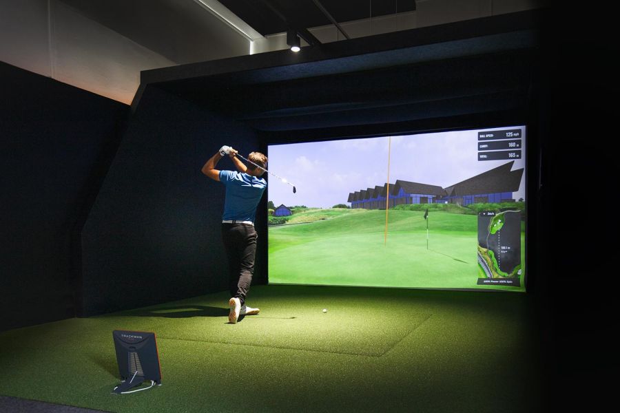 Golfer swinging at a tee in a dark indoor simulator bay with green turf